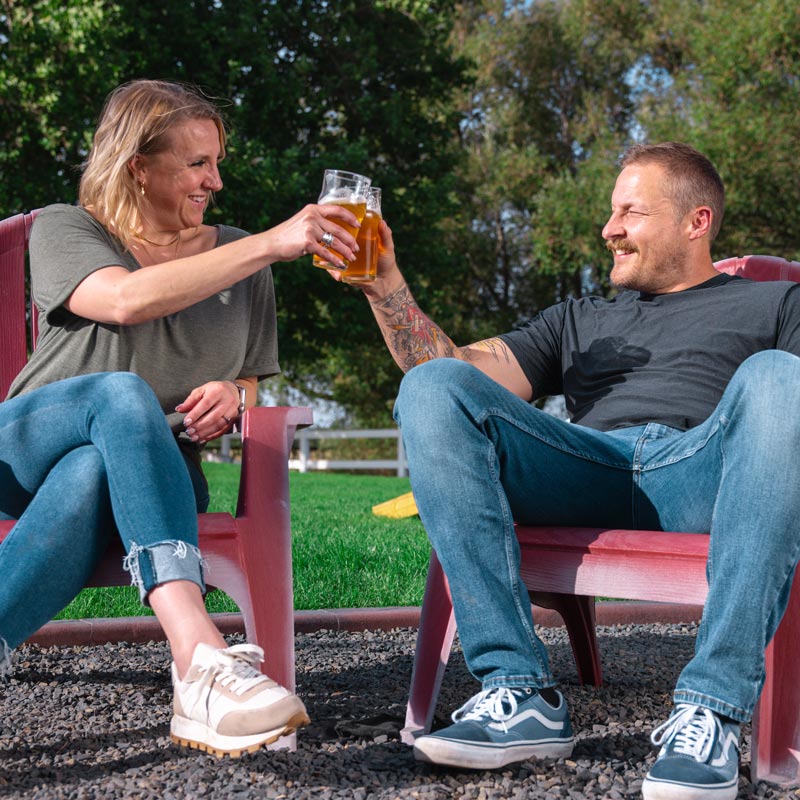 A smiling couple sitting outside on lawn chairs and clinking beer glasses together.